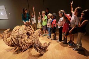 A group of school children gather around a sculpture made of wood during an educational program at the UM Museum
