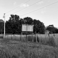 Black and white photo of the grassy side of the road with an empty road sign and a fence.